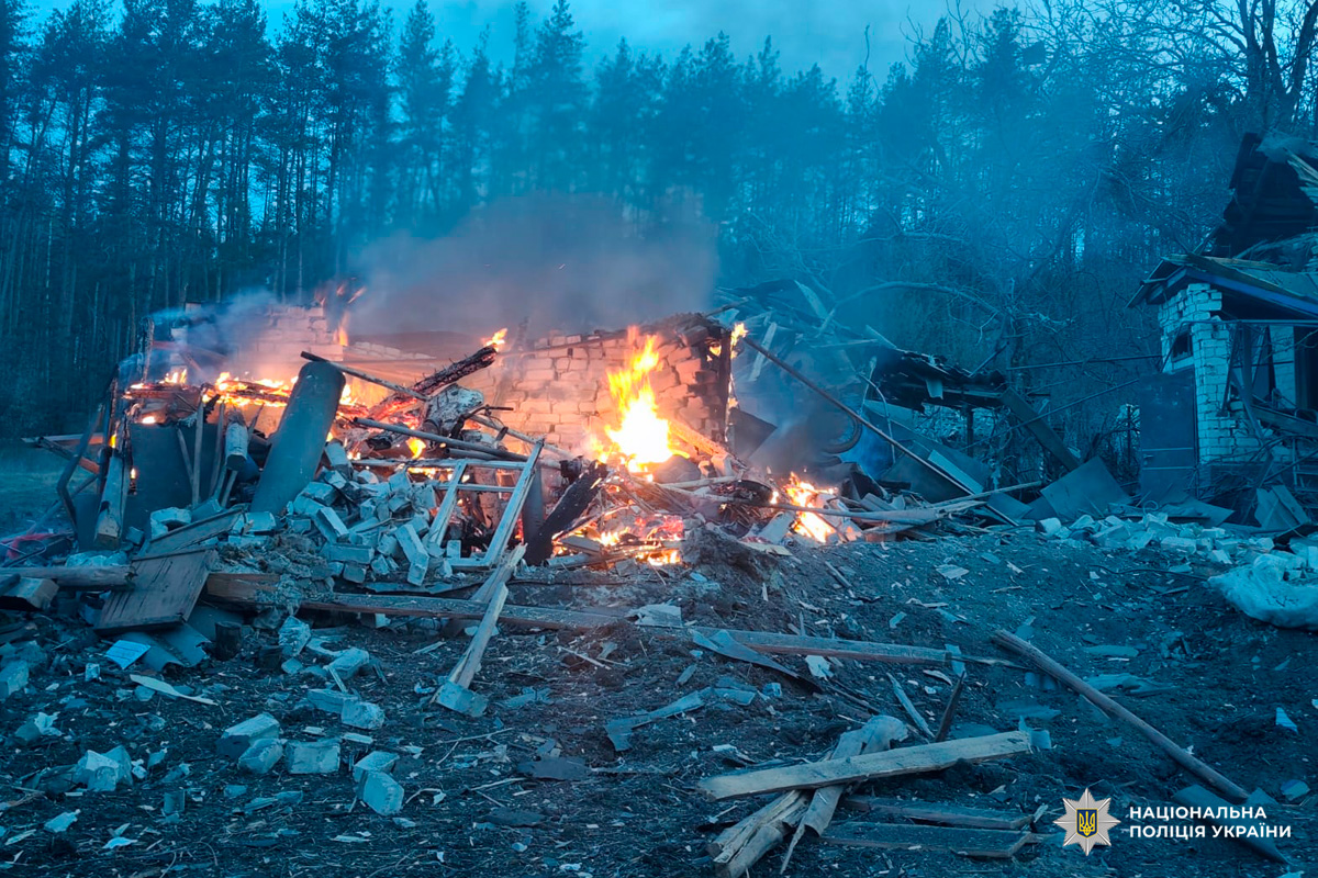 Будівля, зруйнована та охоплена вогнем, уламки цегли й дерева розкидані навколо.