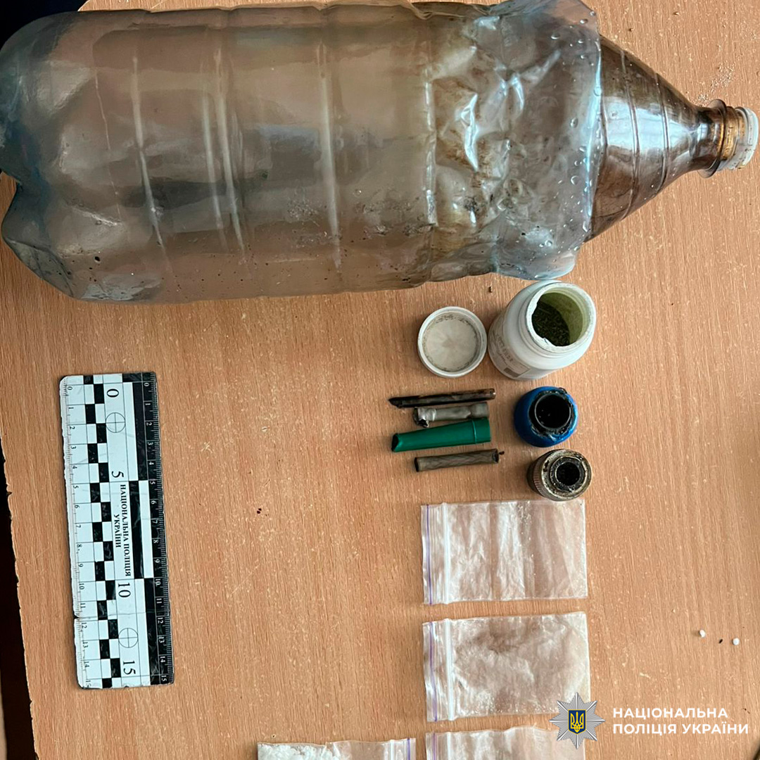 На дерев’яній поверхні викладені вилучені предмети: використана пластикова пляшка, кілька контейнерів із порошком, невеликі циліндричні об’єкти, скляні баночки та прозорі пакети. Ліворуч розташована вимірювальна шкала.