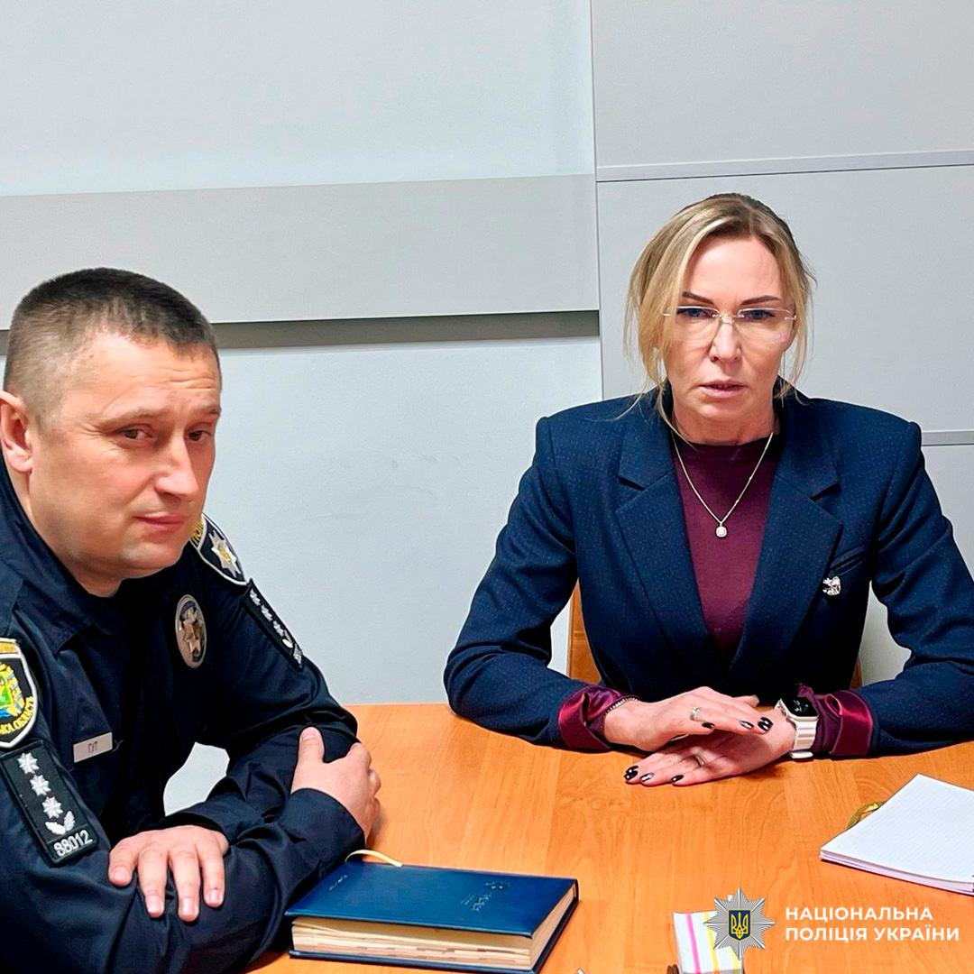 Двоє учасників офіційної зустрічі сидять за дерев’яним столом. Один у формі Національної поліції України, інший у діловому одязі. На столі книга з закладкою, блокнот та документи.