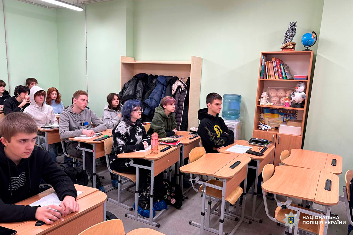 Учні сидять за дерев’яними партами у класі зі світло‑зеленими стінами. На задньому плані книжкова шафа з книгами, іграшками, глобусом та декоративною совою, а також кулер для води.