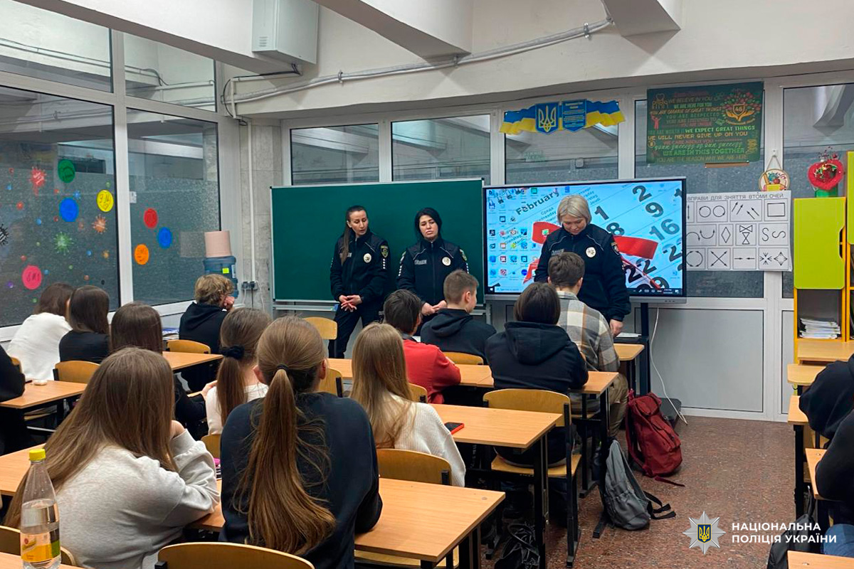 У класі троє офіцерів Національної поліції України у формі стоять перед дошкою та цифровим екраном, звертаючись до учнів. Діти сидять за партами й уважно слухають.