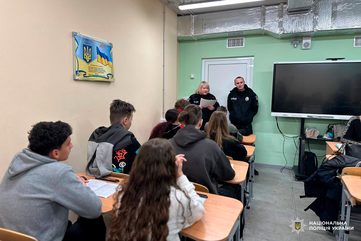 Учні сидять за партами у класі з зеленими стінами. Перед ними двоє офіцерів Національної поліції України у формі, один тримає аркуш і виступає. На стіні великий монітор та табличка з Державним Гімном України й символікою.