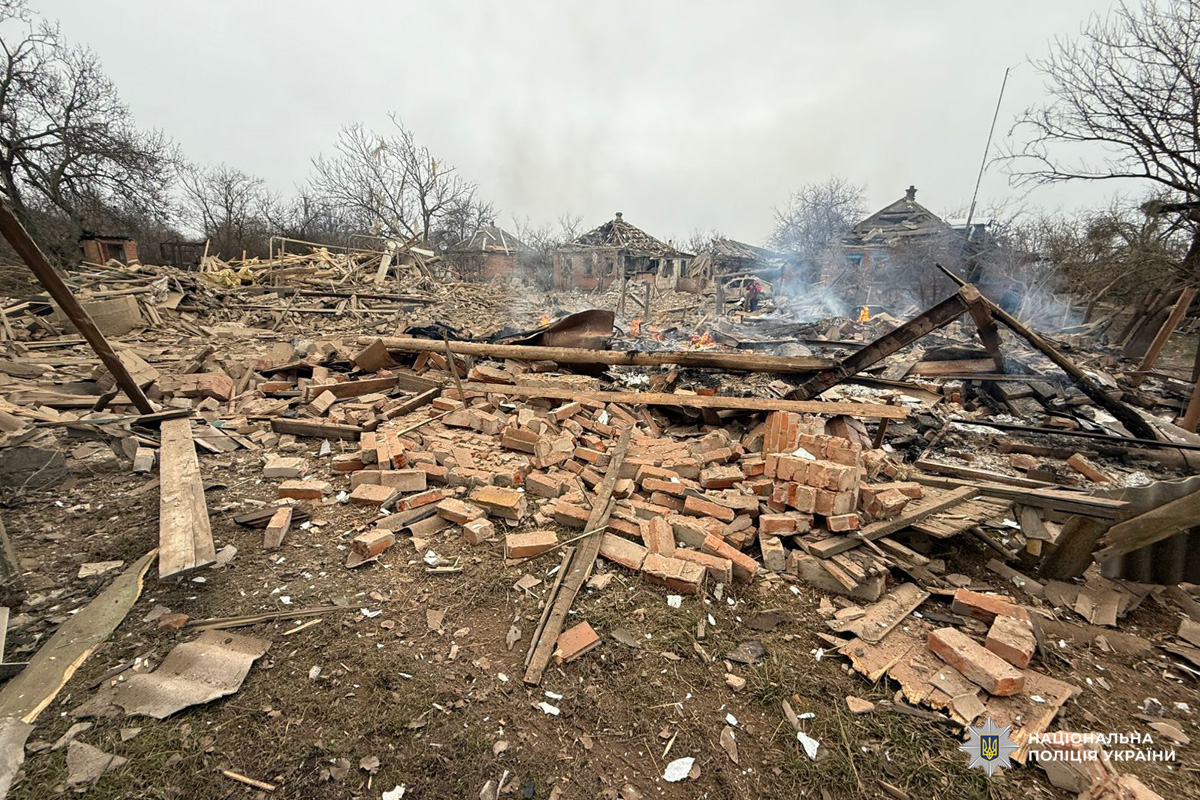 На передньому плані уламки цегли та деревини, у глибині пошкоджені будівлі з осередками пожежі.