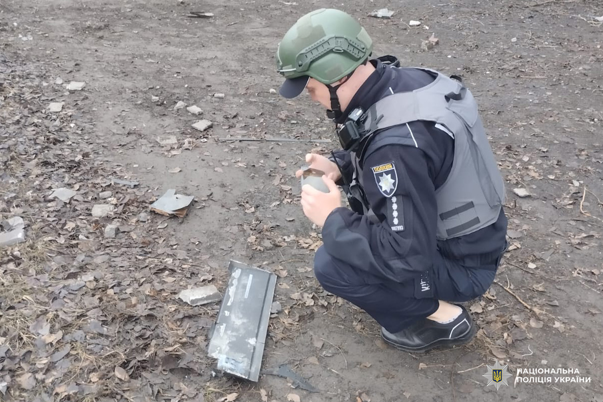 Поліцейський у захисному спорядженні оглядає металевий предмет серед уламків після вибуху.