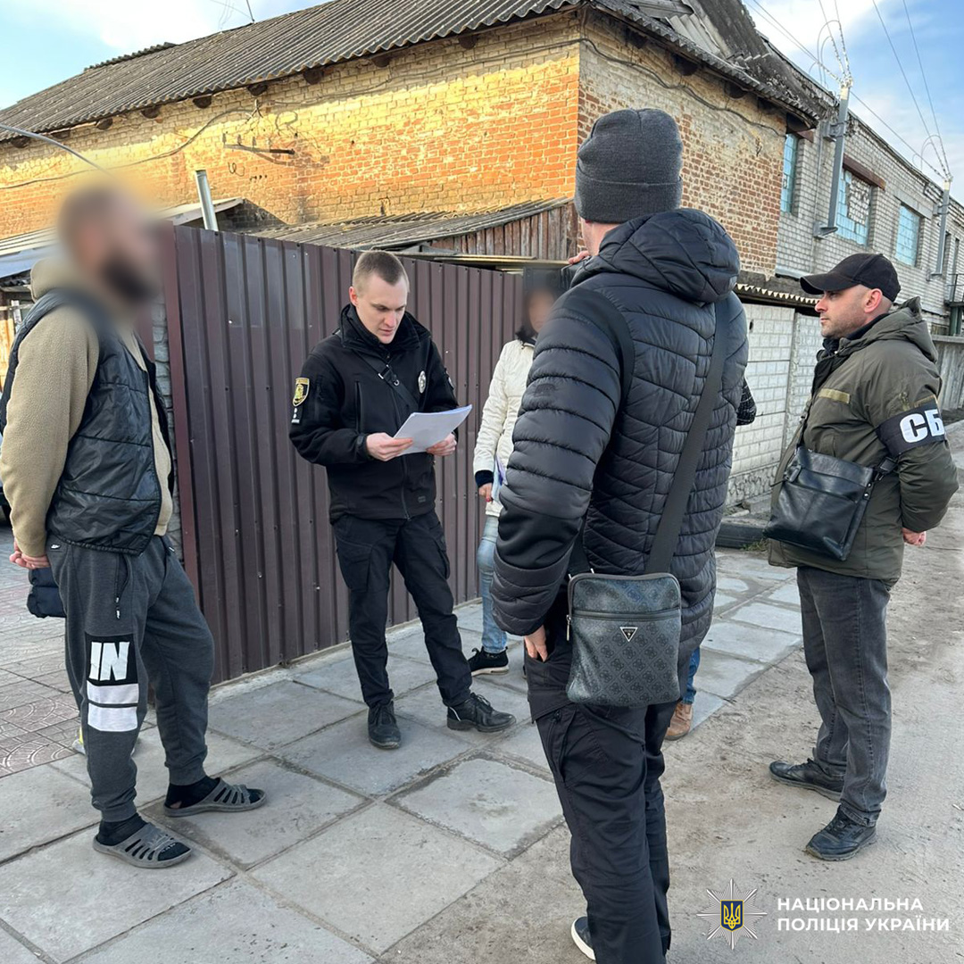 Біля паркану та старих будівель поліцейський читає документ, поруч стоять інші учасники операції, серед них людина з пов’язкою СБУ.