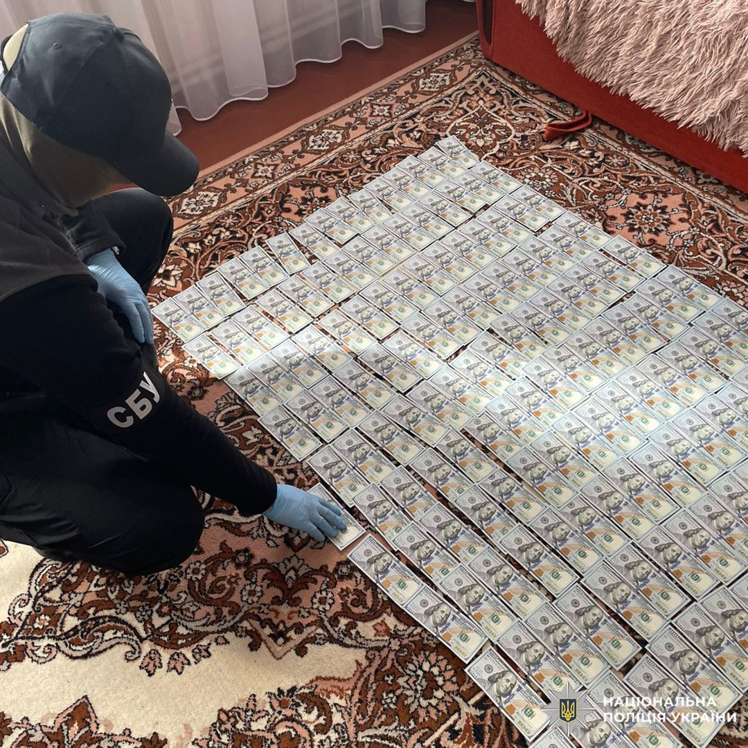 На килимі розкладені сотні купюр у доларах США, співробітник СБУ у темному одязі фіксує знахідку.