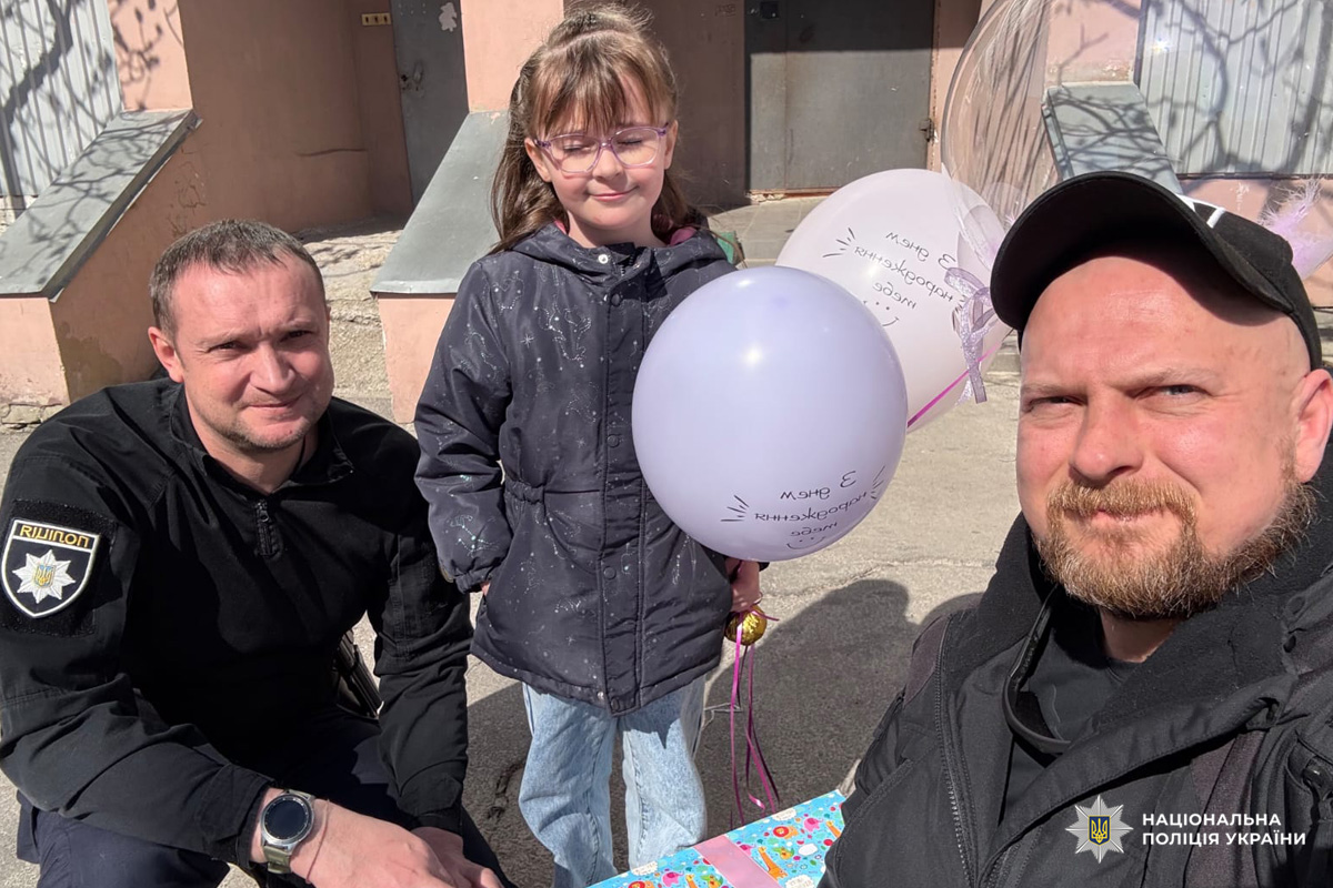 Поліцейські у формі вітають дитину з днем народження, дитина тримає повітряні кульки з написом українською.