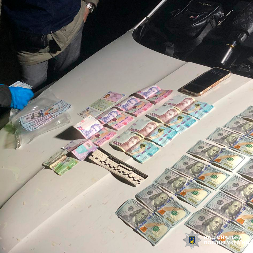 Вилучені кошти різних валют задокументовані на капоті автомобіля, поруч — речові докази та поліцейські матеріали.