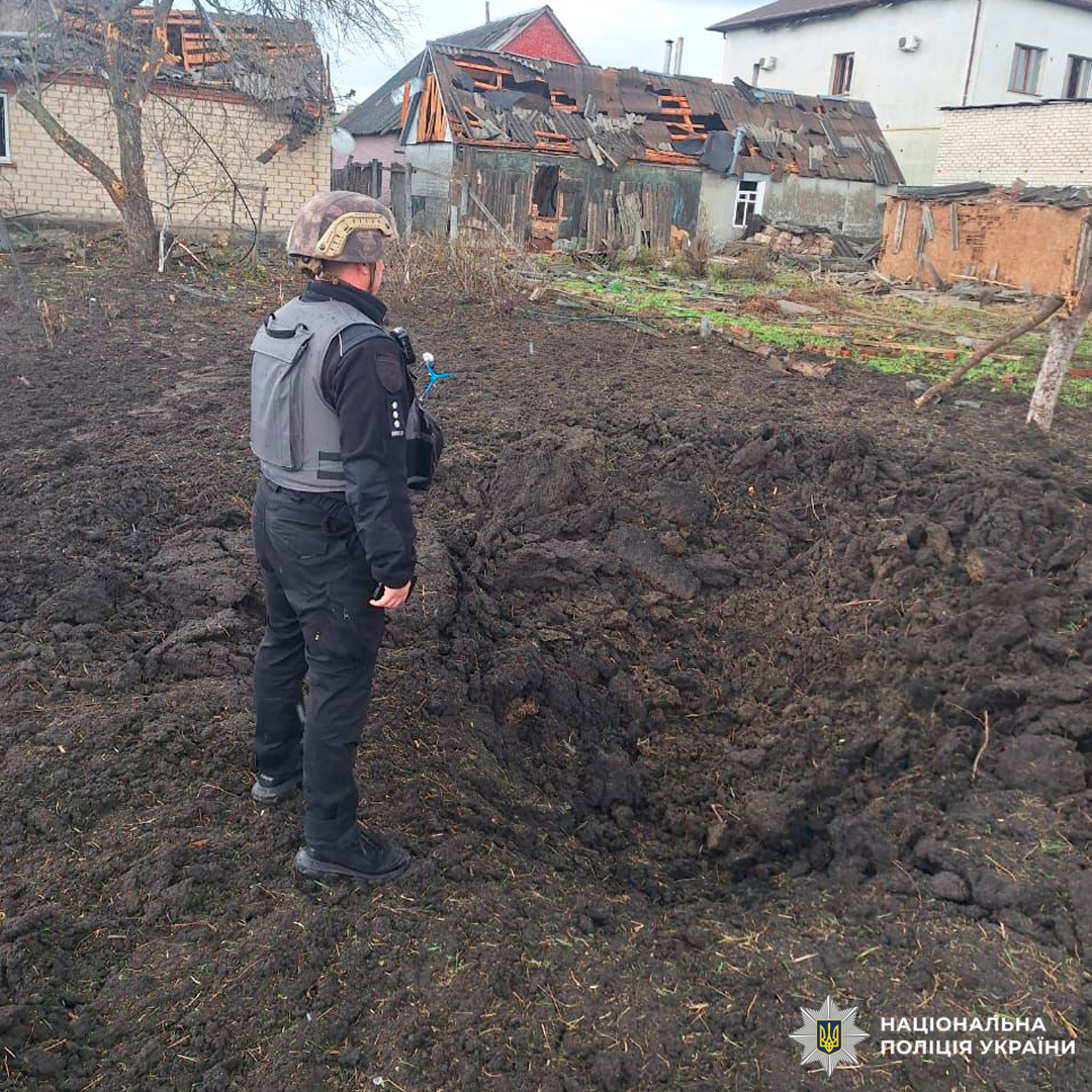 Поліцейський оглядає вирву від вибуху на подвір’ї приватного будинку з пошкодженими дахами та стінами.