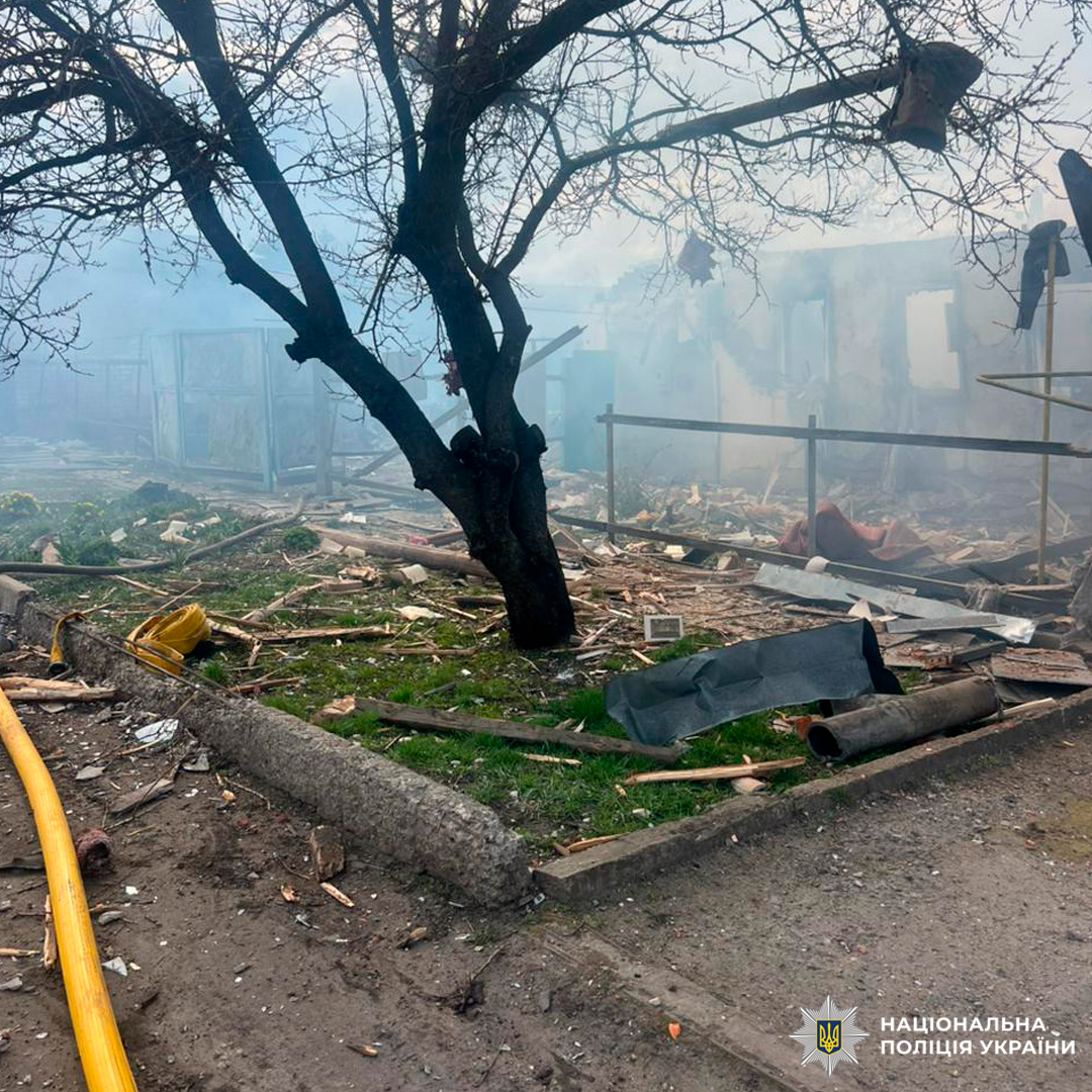 На передньому плані голе дерево серед уламків деревини, металу та сміття; позаду будівля з частково зруйнованими стінами, крізь дим чи туман видно пошкодження; на землі жовтий шланг біля бордюру.