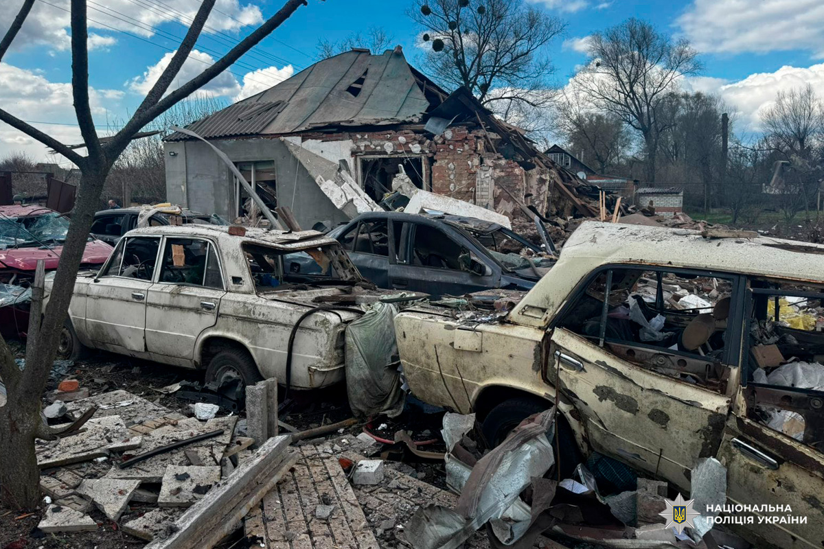 Кілька сильно пошкоджених і обгорілих автомобілів серед уламків, цегли та металу; позаду будинок із частково обваленим дахом і стінами; навколо голі дерева, похмуре небо.