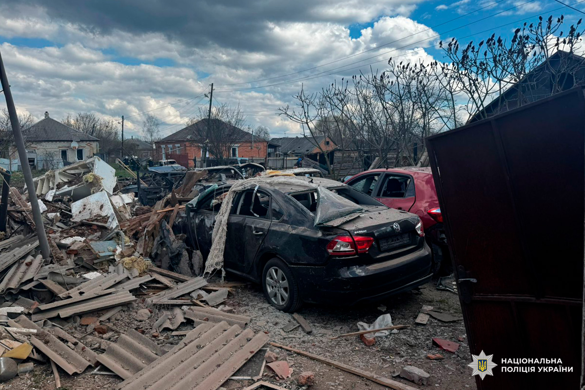Пошкоджені автомобілі, серед них чорний Volkswagen і червоний легковик, навколо уламки дахових матеріалів, цегла та сміття; позаду частково вцілілі будинки під хмарним небом.