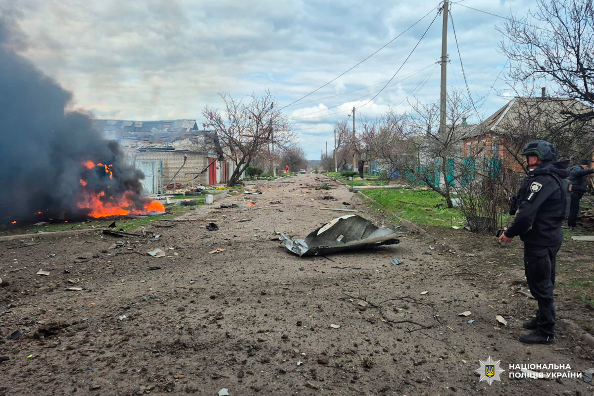 Пожежа біля пошкодженої будівлі, чорний дим здіймається над вулицею, яка вкрита уламками. Праворуч поліцейський у захисному спорядженні оглядає територію.