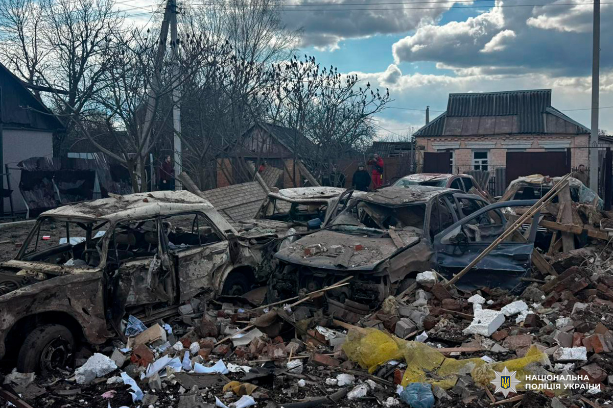 Зруйновані транспортні засоби серед цегли та уламків, пошкоджені житлові будинки, люди оглядають територію після вибуху.