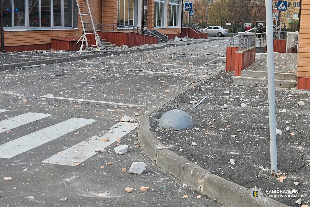 Пошкоджена будівля та прилегла територія після вибуху: уламки бетону, цегли й металу розкидані на тротуарі та дорозі, зафіксовано руйнування інфраструктури.