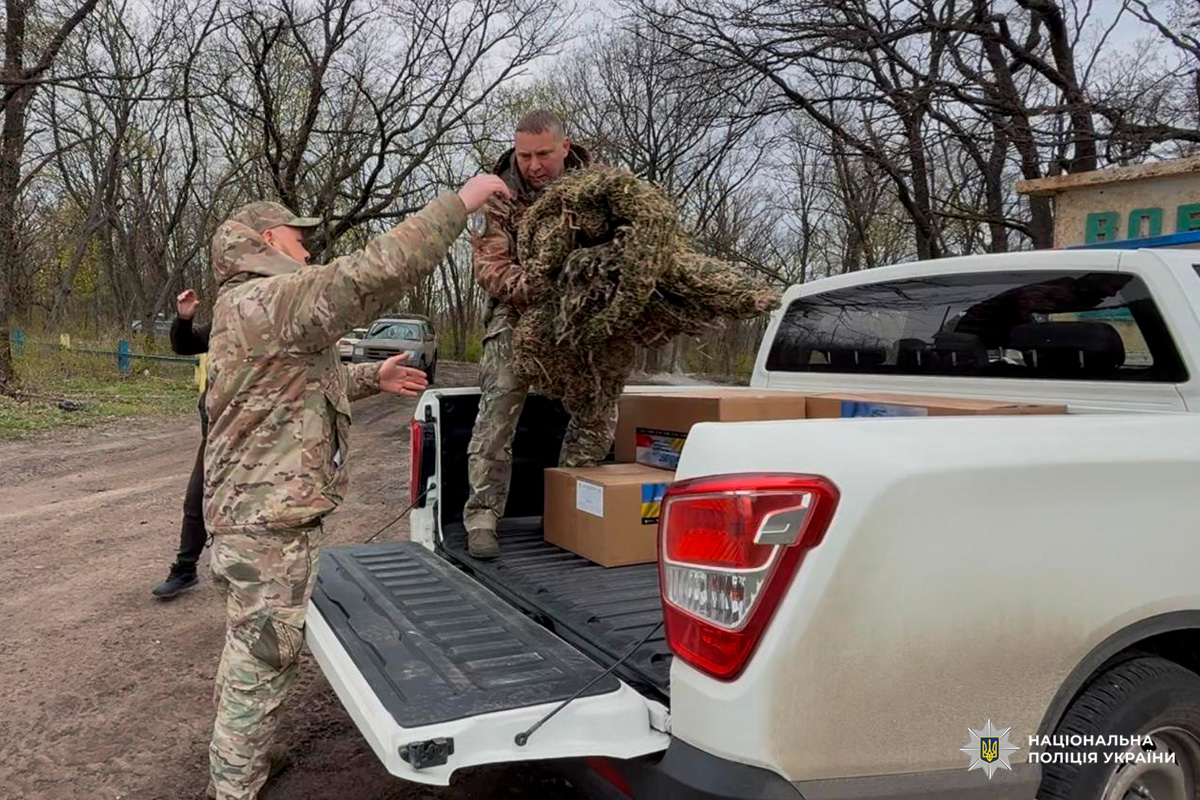 Кілька осіб у камуфляжній формі здійснюють розвантаження припасів із кузова білого пікапа. Один чоловік стоїть у кузові та тримає маскувальне спорядження, інший на землі приймає його. У кузові також видно картонні коробки.
