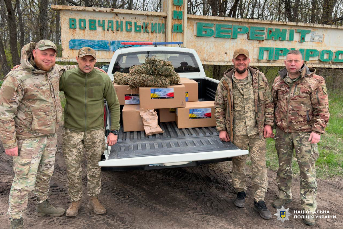 «Чотири особи у камуфляжному та військовому одязі стоять біля білого пікапа. У кузові автомобіля знаходяться картонні коробки та маскувальне спорядження. На задньому плані — великий знак із написами “Вовчанський” та “Бережіть природу”.