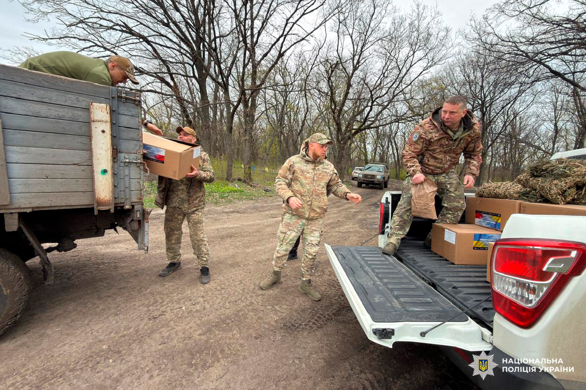 Кілька осіб у камуфляжній формі здійснюють розвантаження картонних коробок із вантажівки та пікапа. Дія відбувається на ґрунтовій дорозі серед дерев без листя, що свідчить про прохолодний сезон. Учасники процесу передають коробки з кузова транспортних засобів на землю.
