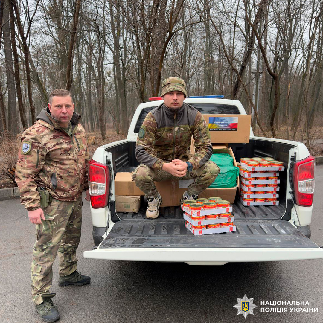 Двоє осіб у камуфляжній формі знаходяться біля білого пікапа. Один стоїть поруч із автомобілем, інший — у кузові, де видно картонні коробки та банки з продуктами. Дія відбувається на відкритій місцевості серед дерев без листя, що свідчить про холодний сезон.