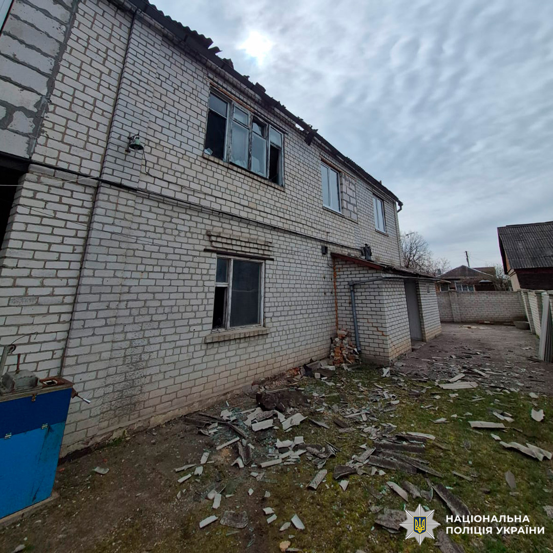 Двоповерхова цегляна будівля з пошкодженим дахом та вибитими вікнами, уламки скла й матеріалів розкидані біля фасаду.
