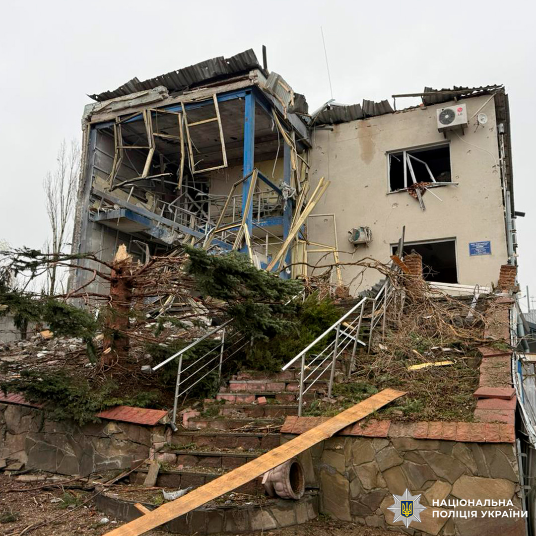 Багатоповерхова будівля з частково обваленими верхніми поверхами, пошкодженими стінами та розбитими вікнами; навколо — уламки та зруйнована рослинність.