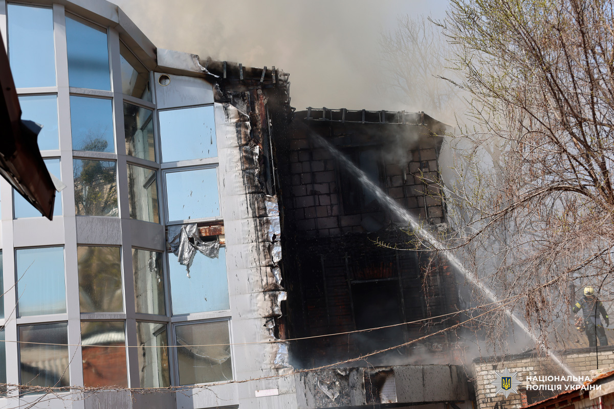 Будівля після пожежі: права частина обгоріла, з неї підіймається дим. Пожежник у захисному спорядженні здійснює гасіння струменем води.