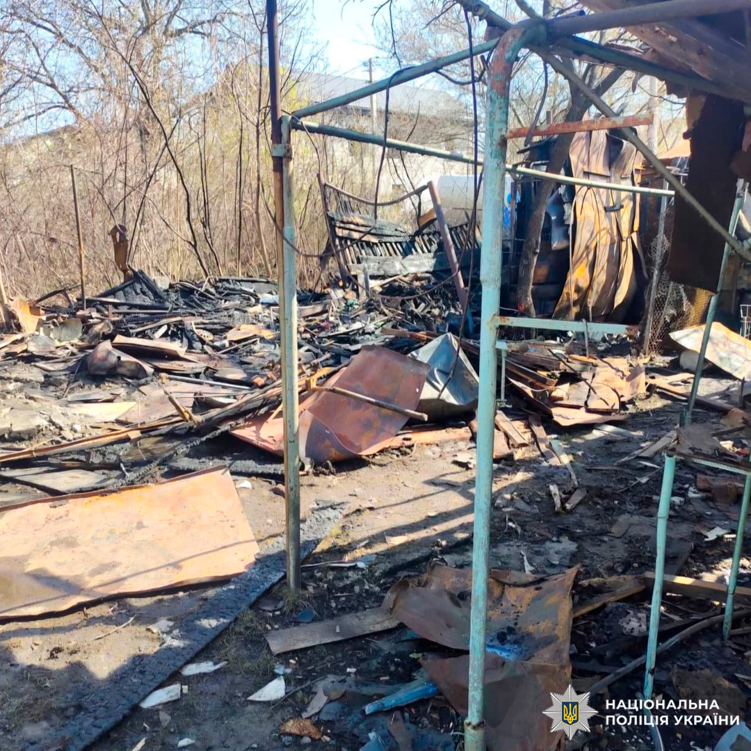 Зруйнована господарська споруда після пожежі: обгорілі дерев’яні балки, деформовані металеві листи та залишки каркасу. На тлі — пошкоджена огорожа й дерева з рідким листям.