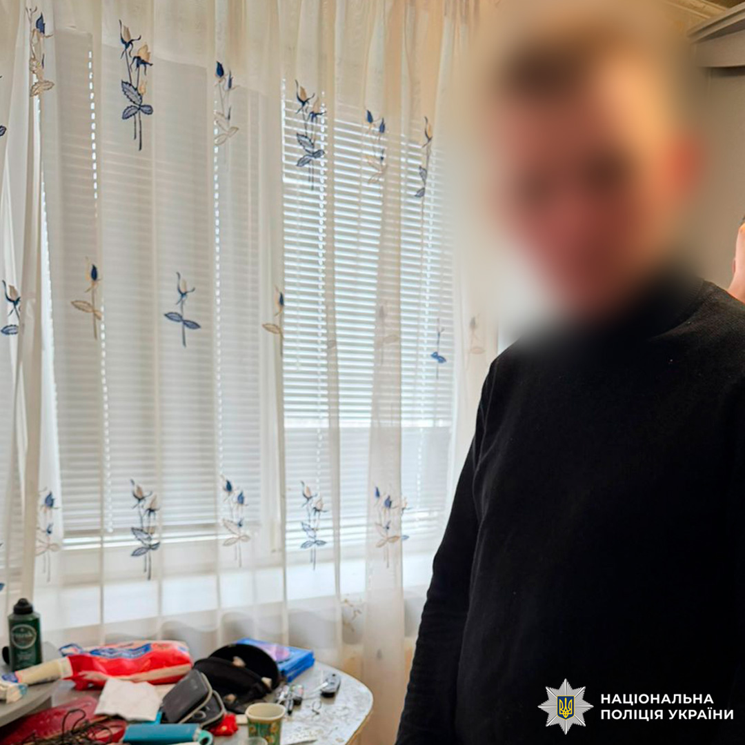 Особа стоїть біля столу з побутовими предметами у кімнаті з вікном та фіранками.