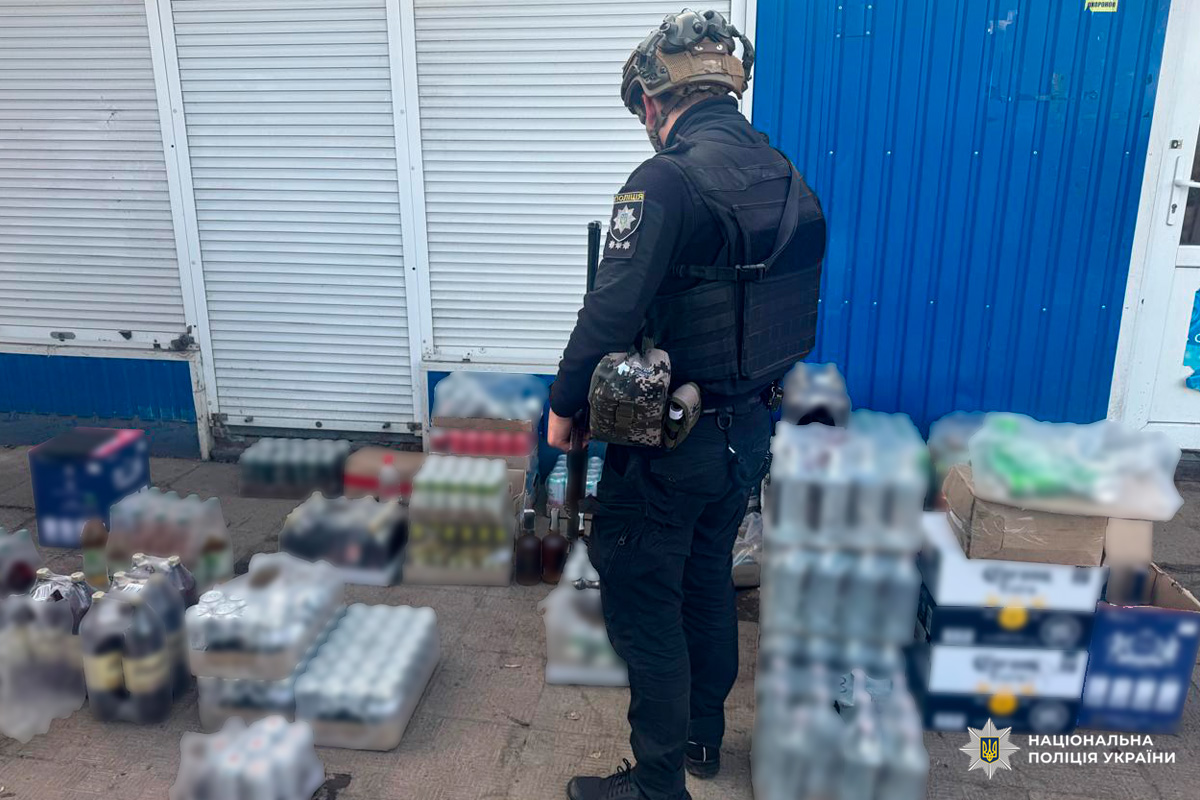 Поліцейський у тактичному спорядженні оглядає вилучені коробки та пляшки напоїв біля будівлі.
