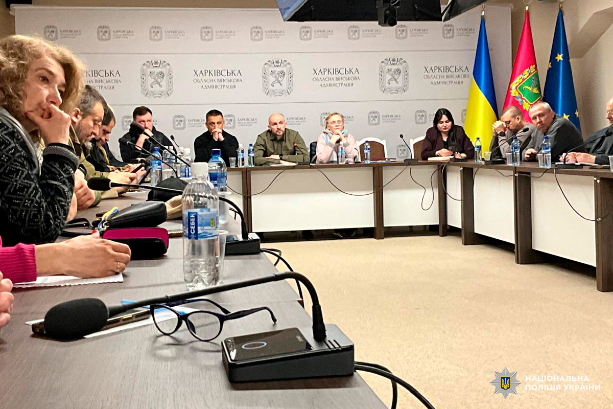 Офіційна нарада у Харківській обласній військовій адміністрації. Учасники сидять за столом із мікрофонами та документами, позаду — прапори України, регіону та ЄС.