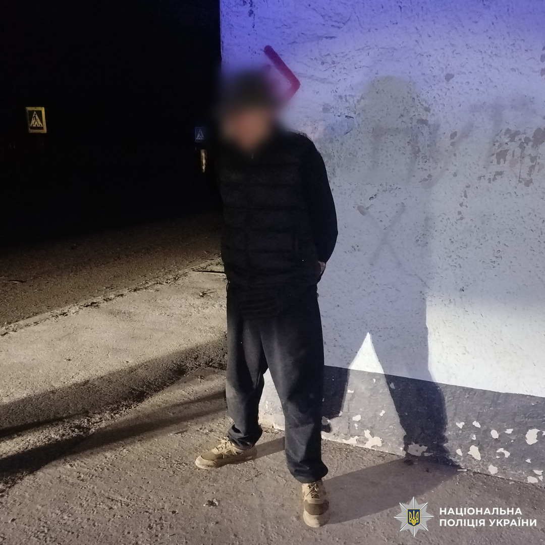 Чоловік стоїть біля стіни будівлі у темний час доби. Обличчя розмите. Він одягнений у темний одяг — куртку та штани, руки тримає за спиною. На стіні поруч видно дорожній знак пішохідного переходу.