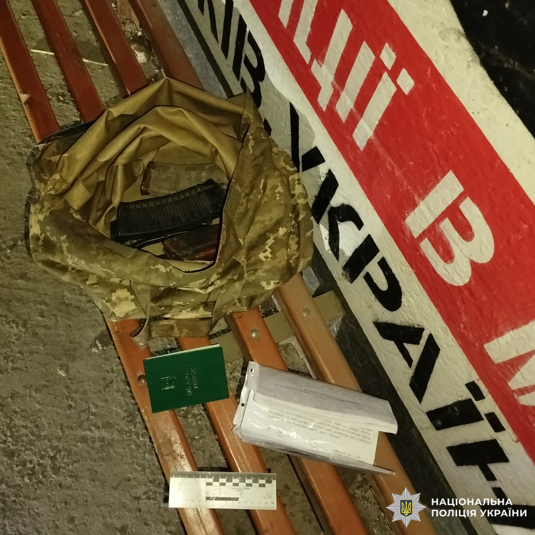 На лавці лежить камуфльована сумка, всередині якої видно предмет, схожий на магазин до вогнепальної зброї. Поруч розміщені документи, зелена книжечка (ймовірно посвідчення або паспорт), а також лінійка для фіксації масштабу. Фон — бетонна поверхня та стіна з написом.