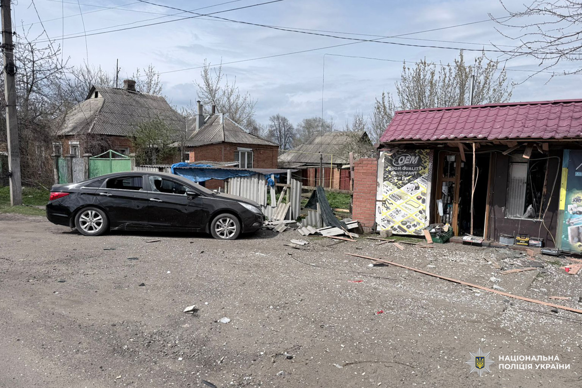 На ґрунтовій дорозі стоїть пошкоджений чорний седан. Поруч будівля з червоним дахом має вибиті вікна та частково обвалену стіну, уламки розкидані навколо.