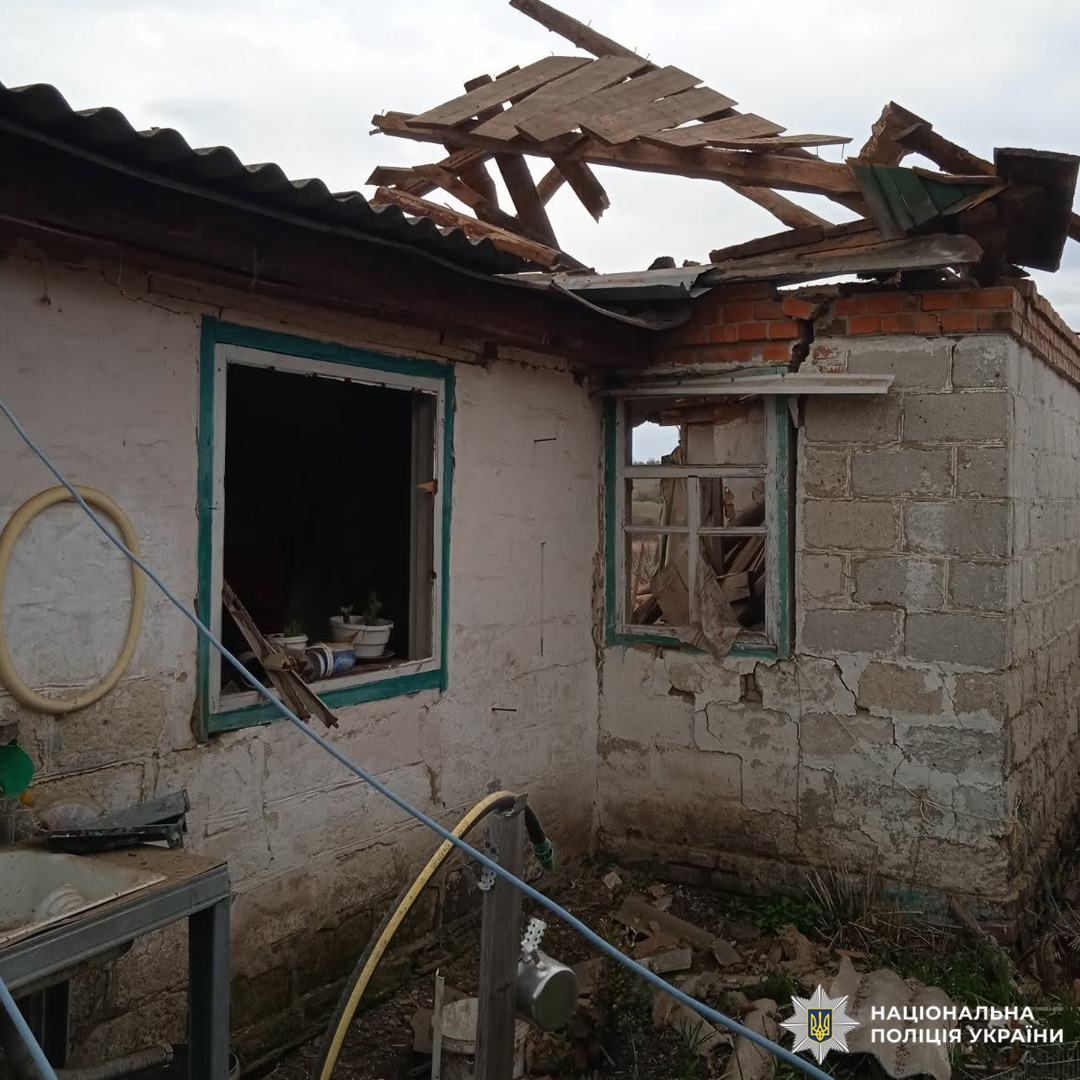 Будівля з частково обваленим дахом, вибитими вікнами та тріщинами в стінах; усередині видно вазони, зовні — господарські предмети.