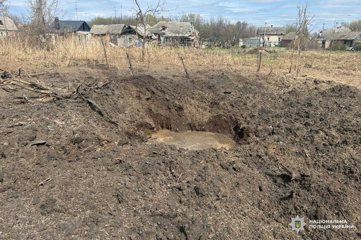 Велика вирва на полі після вибуху, поруч пошкоджені будинки та дерева.