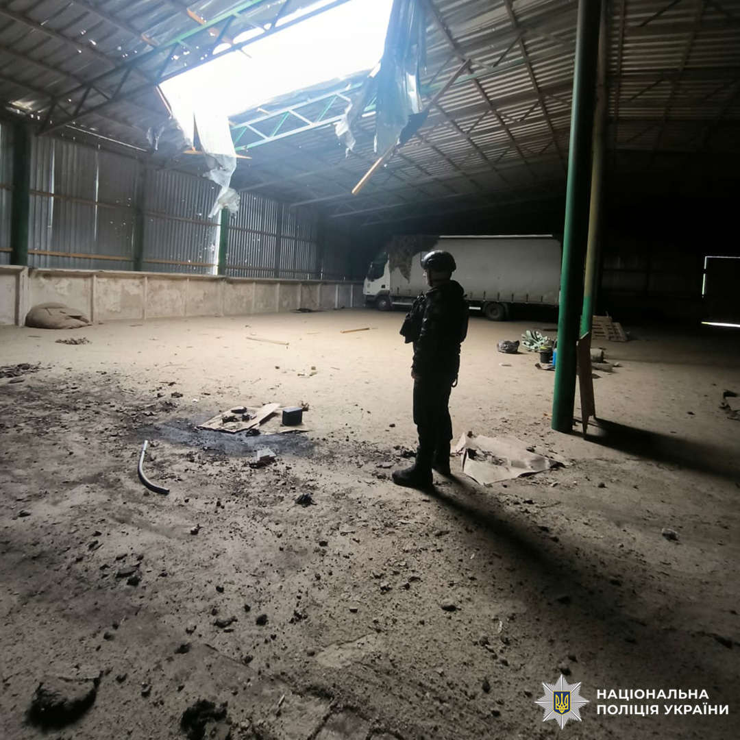 Пошкоджений склад із пробитим дахом, уламками та вантажівкою всередині; поліцейський у тактичному спорядженні проводить огляд.