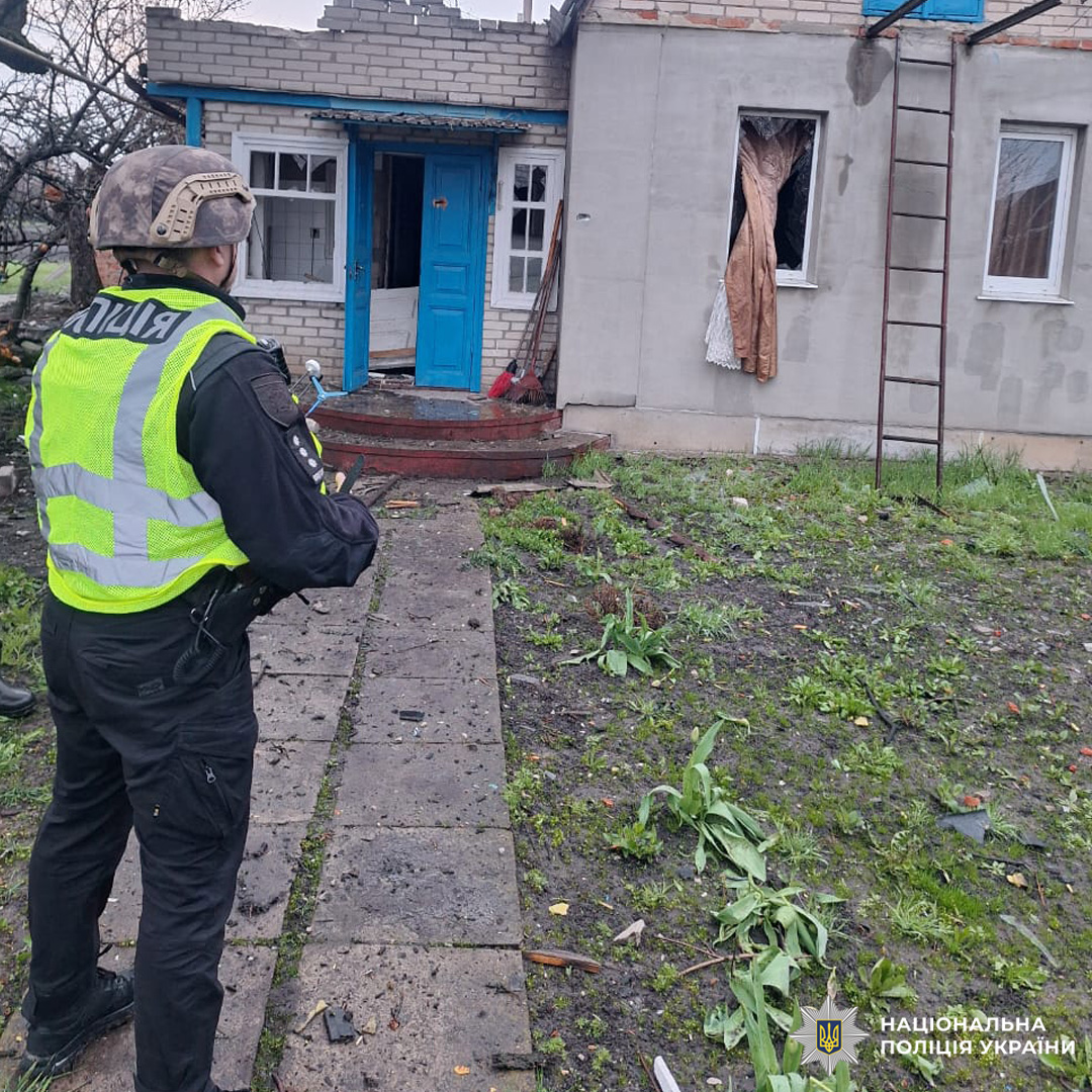 Поліцейський у тактичному спорядженні оглядає пошкоджений житловий будинок із розбитим вікном та розкиданими уламками у дворі.