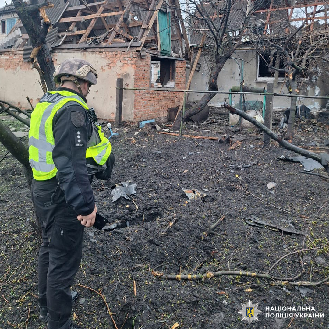 Поліцейський у захисному спорядженні оглядає пошкоджений будинок із частково зруйнованим дахом та вибитими вікнами, серед уламків і вирви на території.