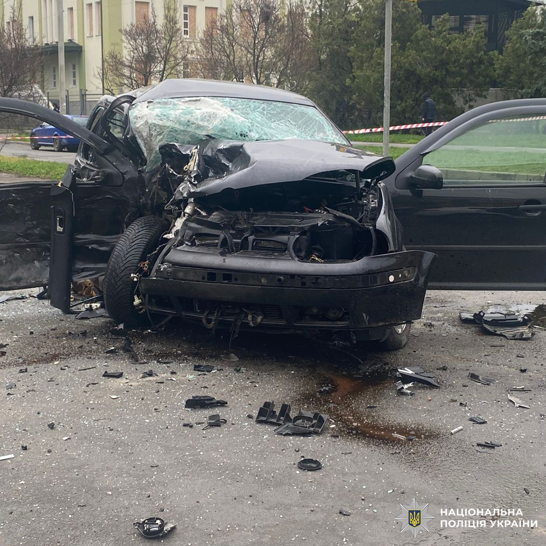 На місці ДТП видно чорний легковий автомобіль із повністю зруйнованим передом, відкритими дверима та уламками скла й деталей на дорозі, територія огороджена стрічкою.