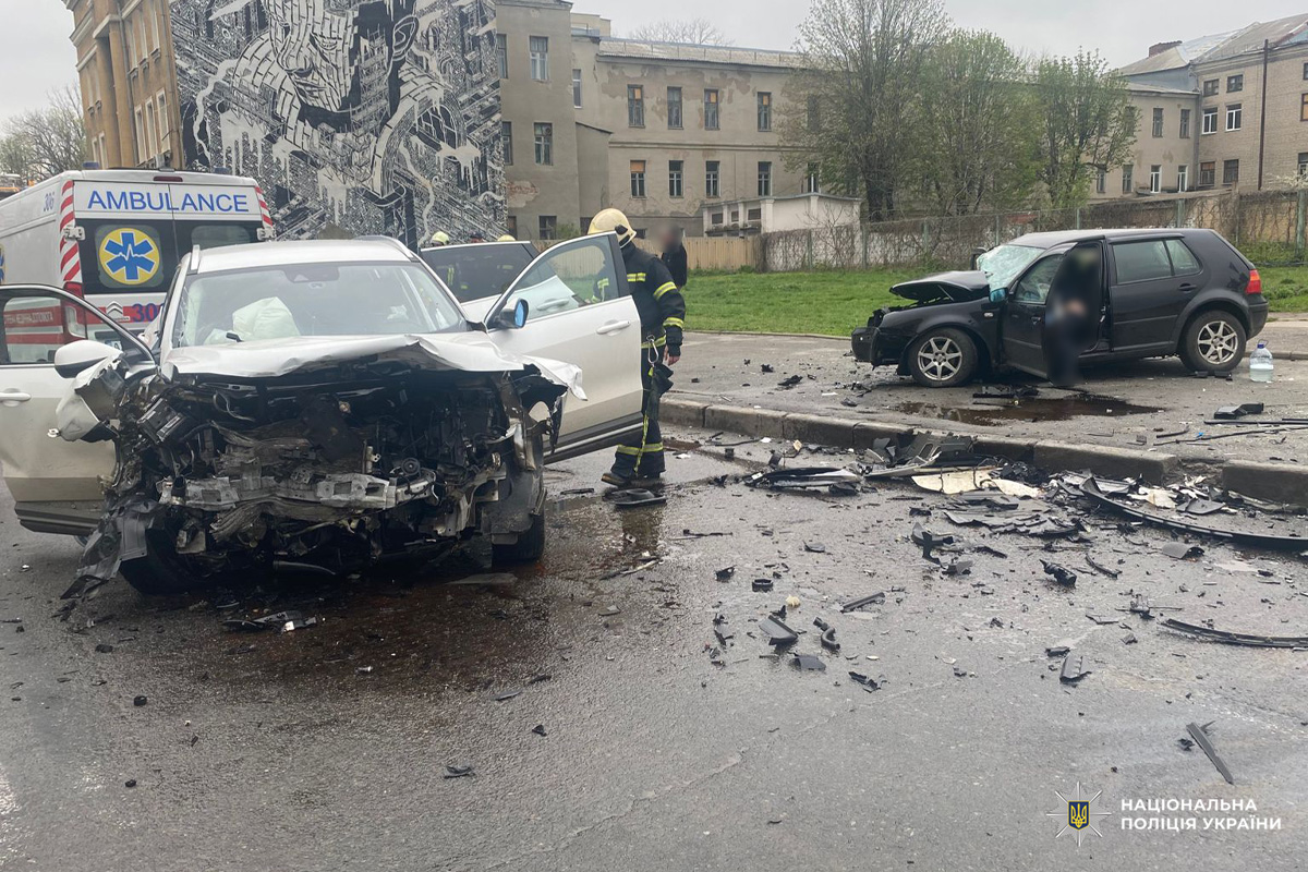 Два пошкоджені автомобілі після зіткнення на міському перехресті, поруч працюють медики та стоїть карета швидкої допомоги.