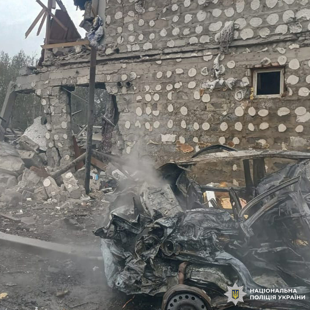 Будівля з обваленим дахом і стінами, перед нею знищений та обгорілий автомобіль серед уламків.