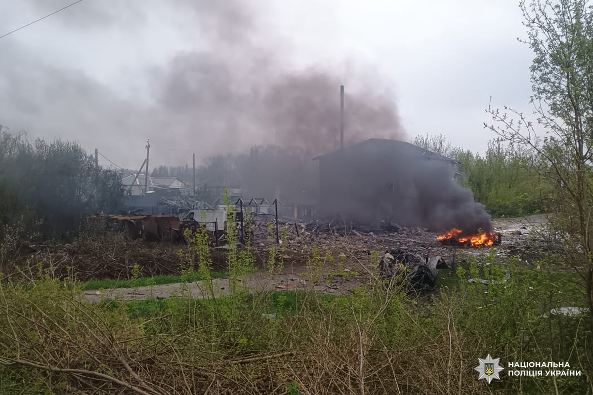 Будівля з пошкодженими стінами, навколо уламки та вогонь, над територією піднімається чорний дим.
