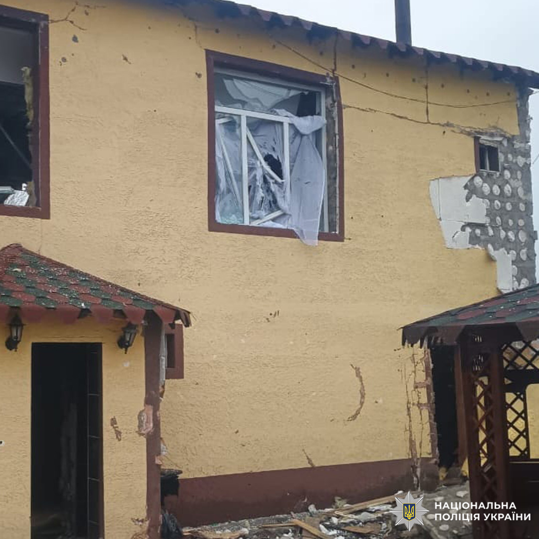 Двоповерховий будинок із жовтим фасадом має тріщини, вибите вікно та пошкоджений дах. На подвір’ї видно уламки та зруйновану дерев’яну конструкцію.