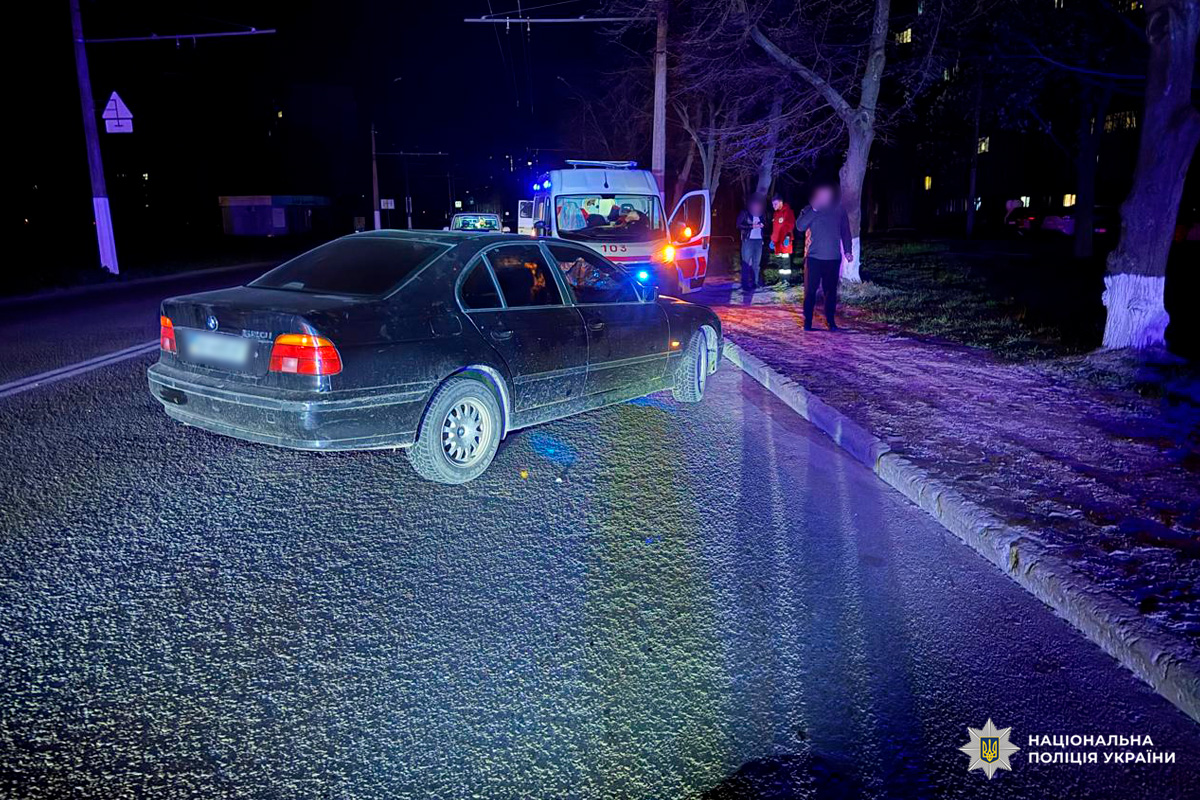 Нічна сцена дорожнього інциденту: чорний седан BMW зупинився частково на дорозі й тротуарі, позаду стоїть карета швидкої допомоги з увімкненими проблисковими маячками та відкритими дверима, навколо кілька людей і рятувальники.