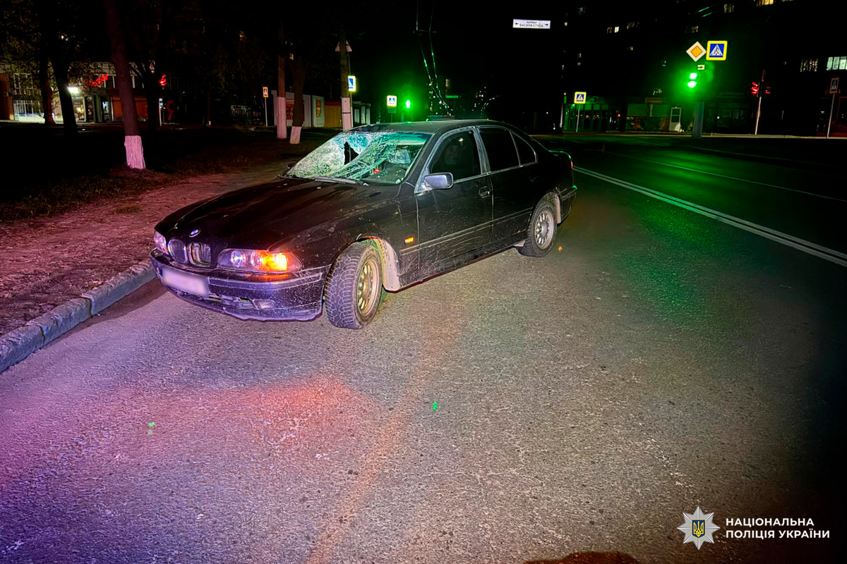 Нічна сцена на міській вулиці: темний седан BMW із розбитим лобовим склом та вм’ятинами на капоті стоїть біля світлофора. Вулицю освітлюють зелені сигнали світлофора та ліхтарі, видно будівлі й дорожні знаки.