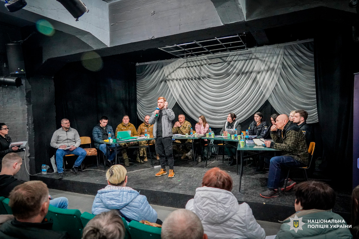 Люди слухають виступ в залі, за столами сидять учасники у формі та цивільному одязі.