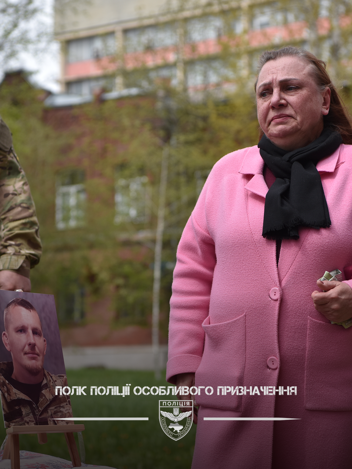Жінка у рожевому пальті та військовослужбовець стоять біля портрета загиблого на вулиці.