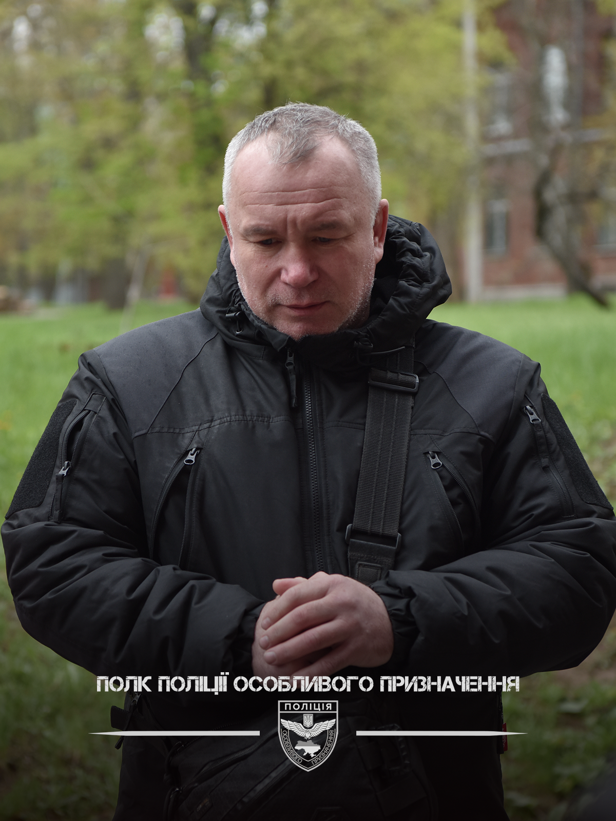 Чоловік у чорній куртці стоїть на подвір’ї біля дерев та будівлі.