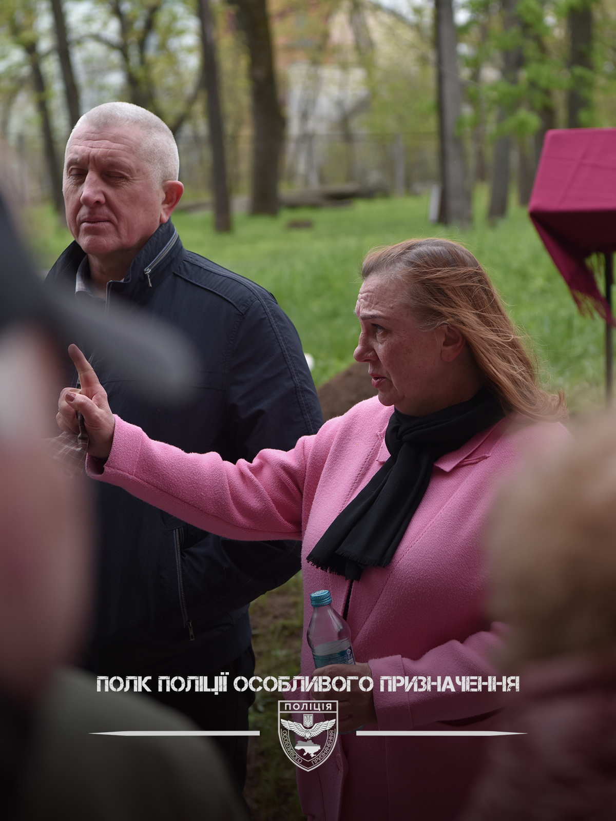 Двоє людей стоять у парку, одна особа в рожевому пальті тримає пляшку води та жестикулює.