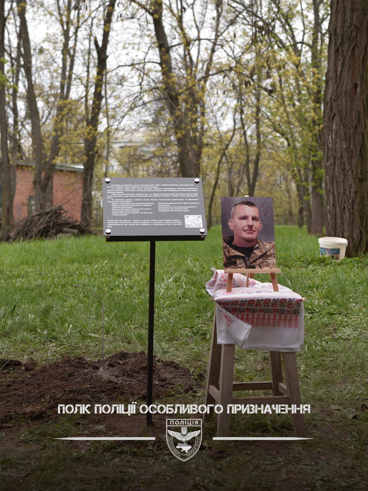 Фото військовослужбовця у рамці на столі з вишитим рушником, поруч пам’ятна плита з текстом та QR‑кодом.